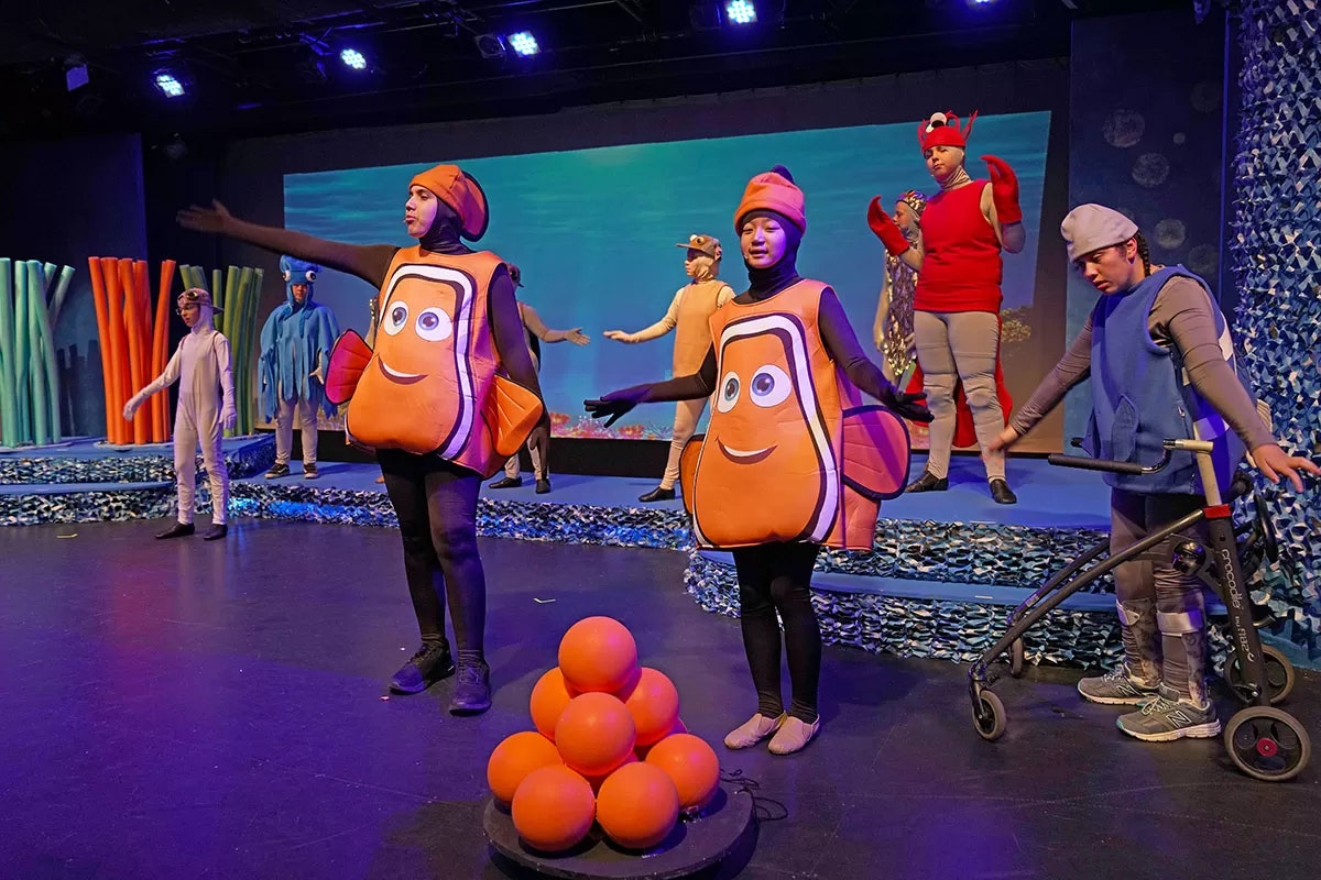 A stage performance with actors in colorful costumes resembling sea creatures, including clownfish and other marine life.