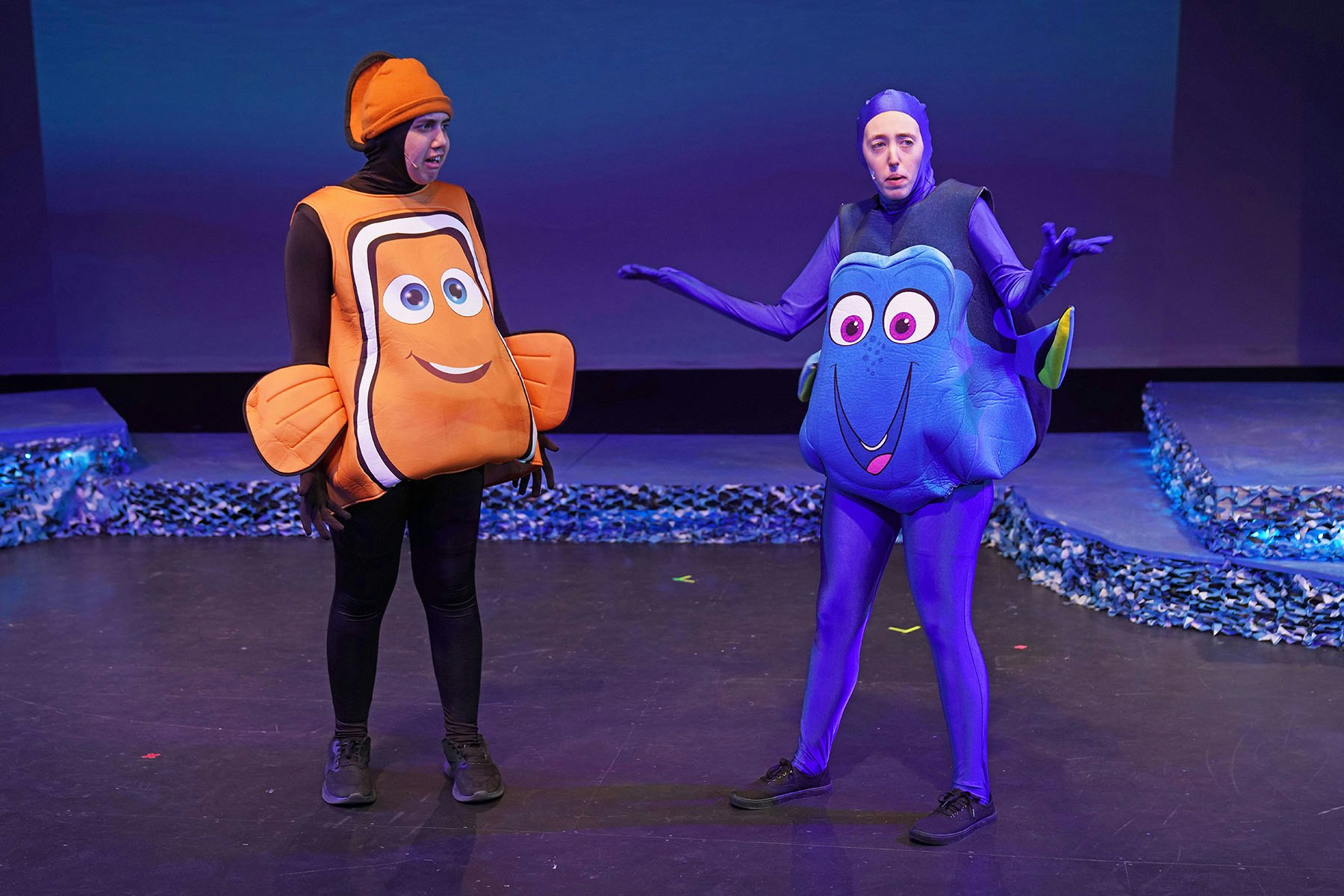 Two people in a play dressed as colorful fish characters on stage.