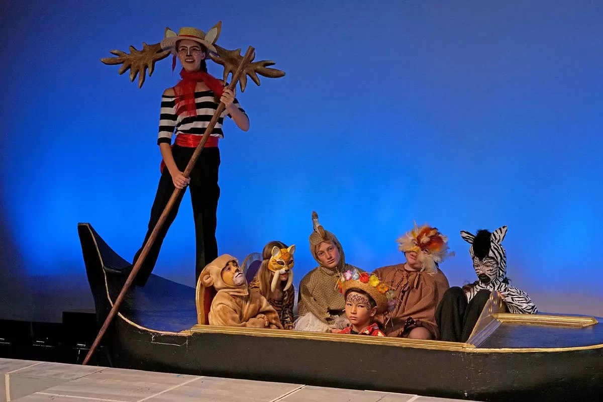 People in animal costumes sitting in a boat on stage, with a performer standing and holding a pole.