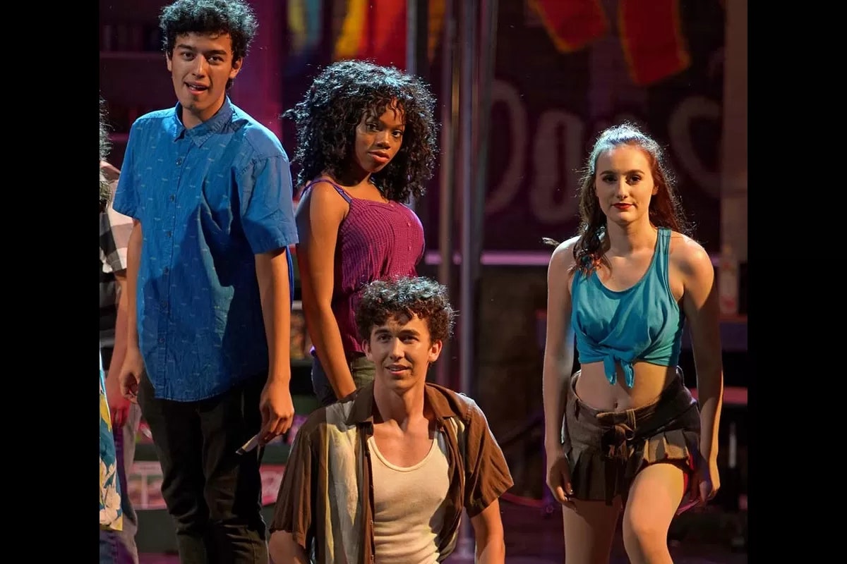 A group of four people on stage, dressed in casual, colorful outfits, with a background of lights and vibrant colors.