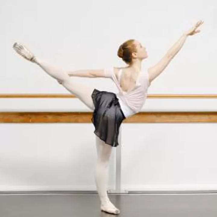 Ballet dancer in extended pose by a barre, wearing a white top, black skirt, and pointe shoes.