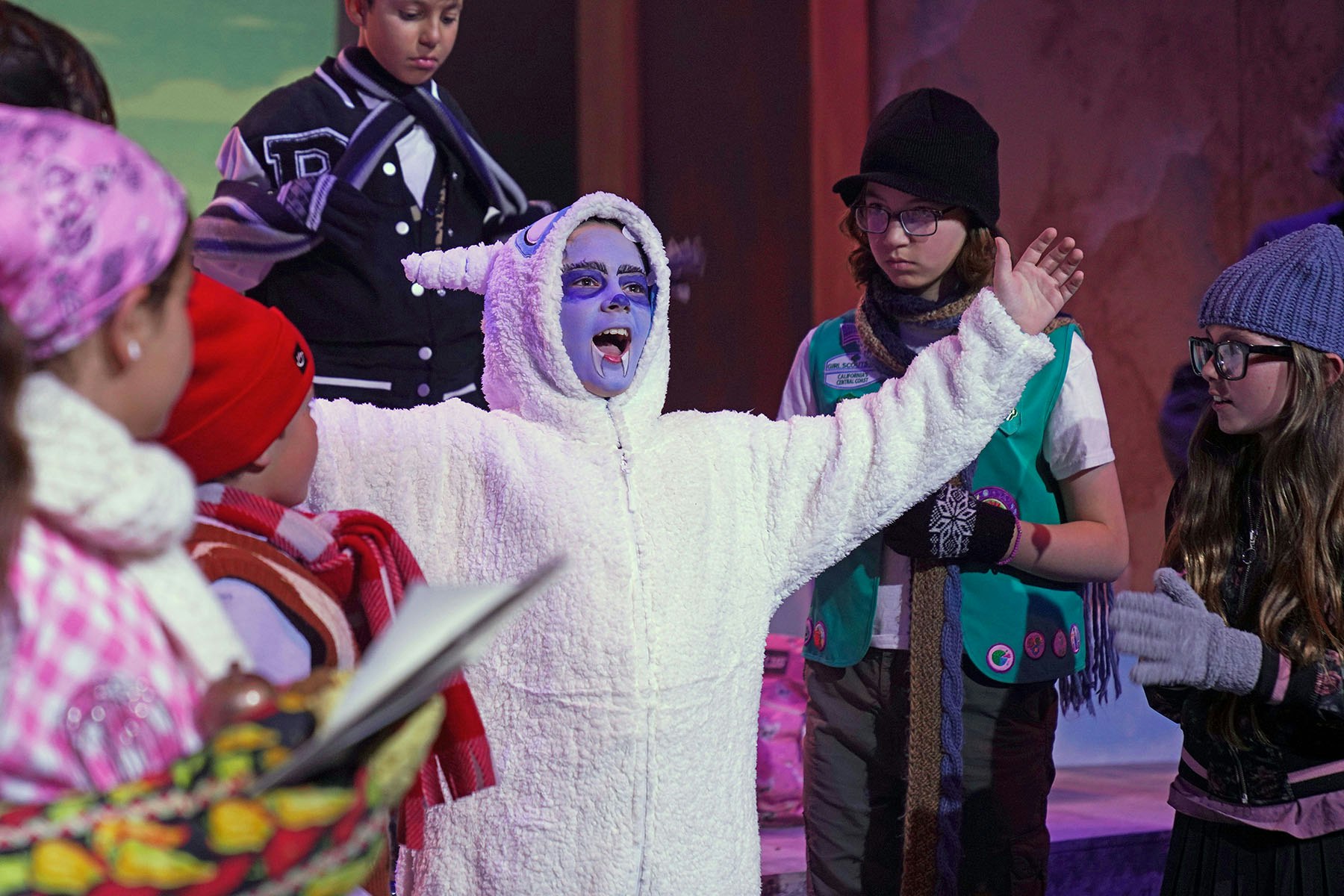 A performer in a white fluffy costume and face paint is expressing excitement in front of an audience of children dressed for winter.