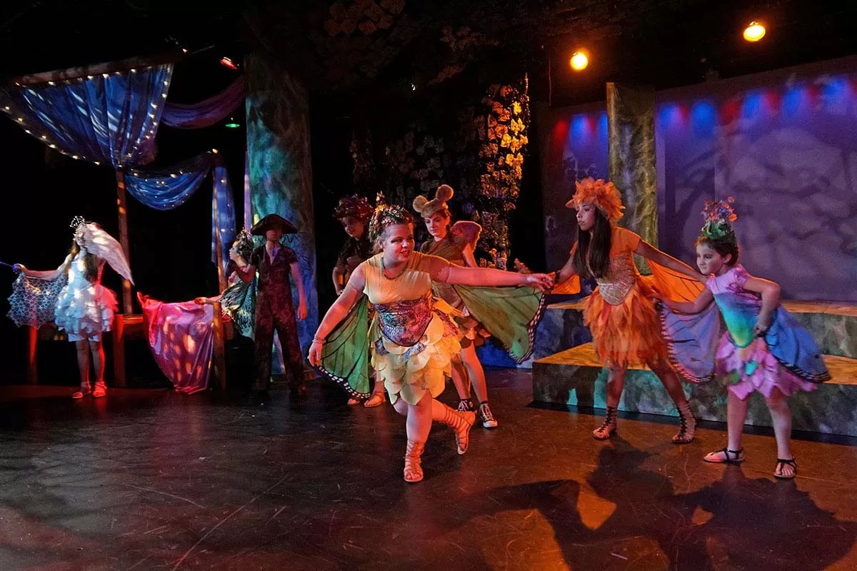 Performers in colorful costumes on stage, under vibrant lighting, resembling a theatrical play or dance performance.