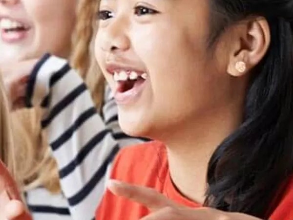 A smiling child with missing teeth, wearing earrings and a red shirt, gesturing with a blurred background.