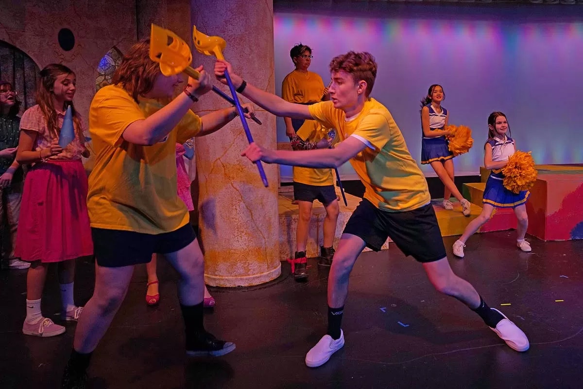 People on stage, some in yellow shirts play-fighting with toy swords, others cheerleading and watching.