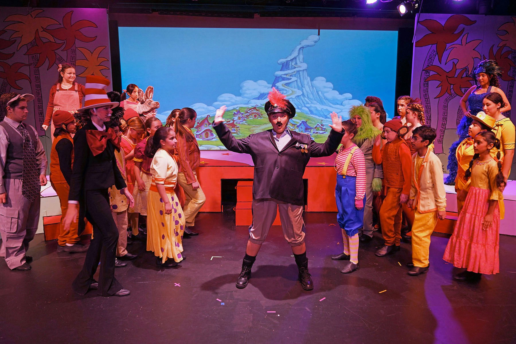 A colorful stage scene featuring children and adults in costumes performing, likely from a musical or theatrical production.