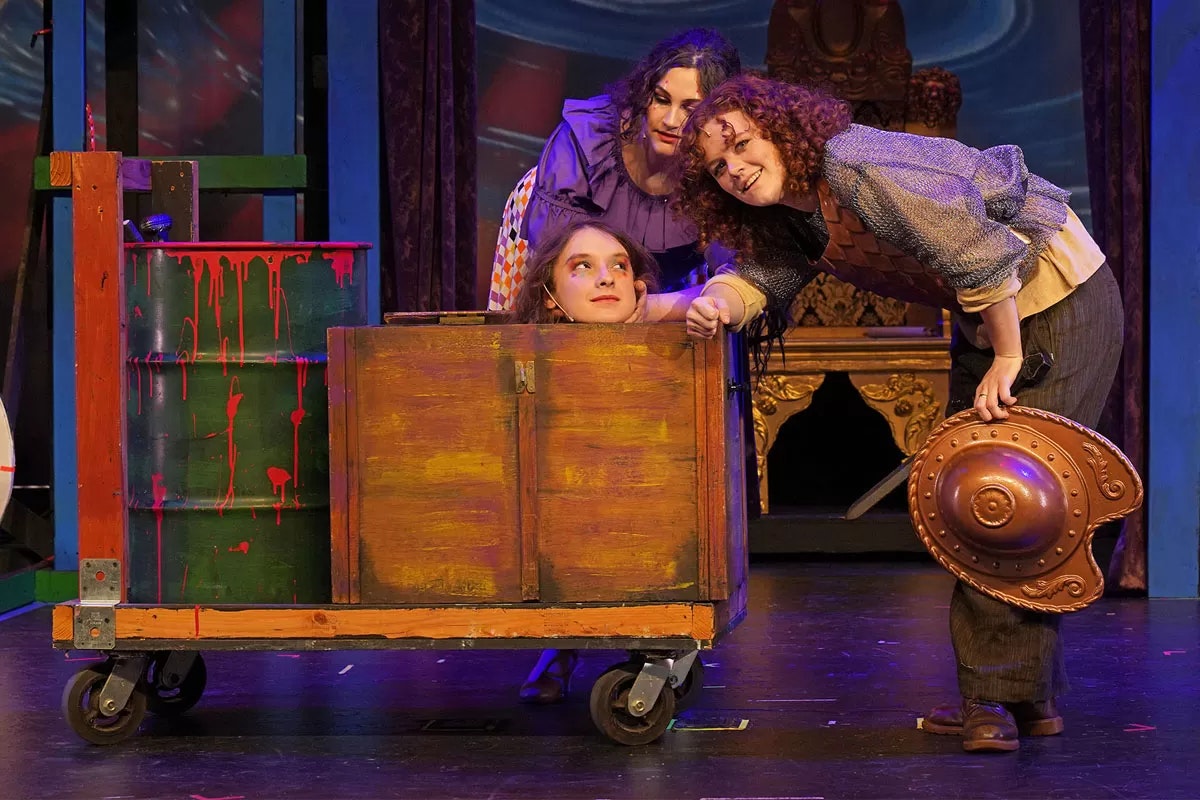 Three people in colorful costumes, one inside a wooden cart, performing a play on stage.