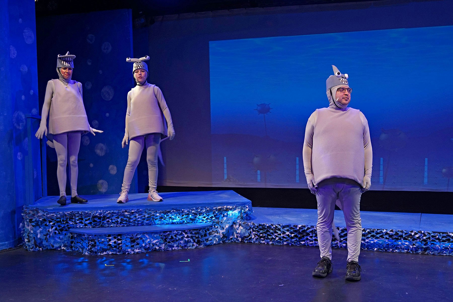 Three people on stage in shark costumes, with an ocean backdrop and blue lighting.