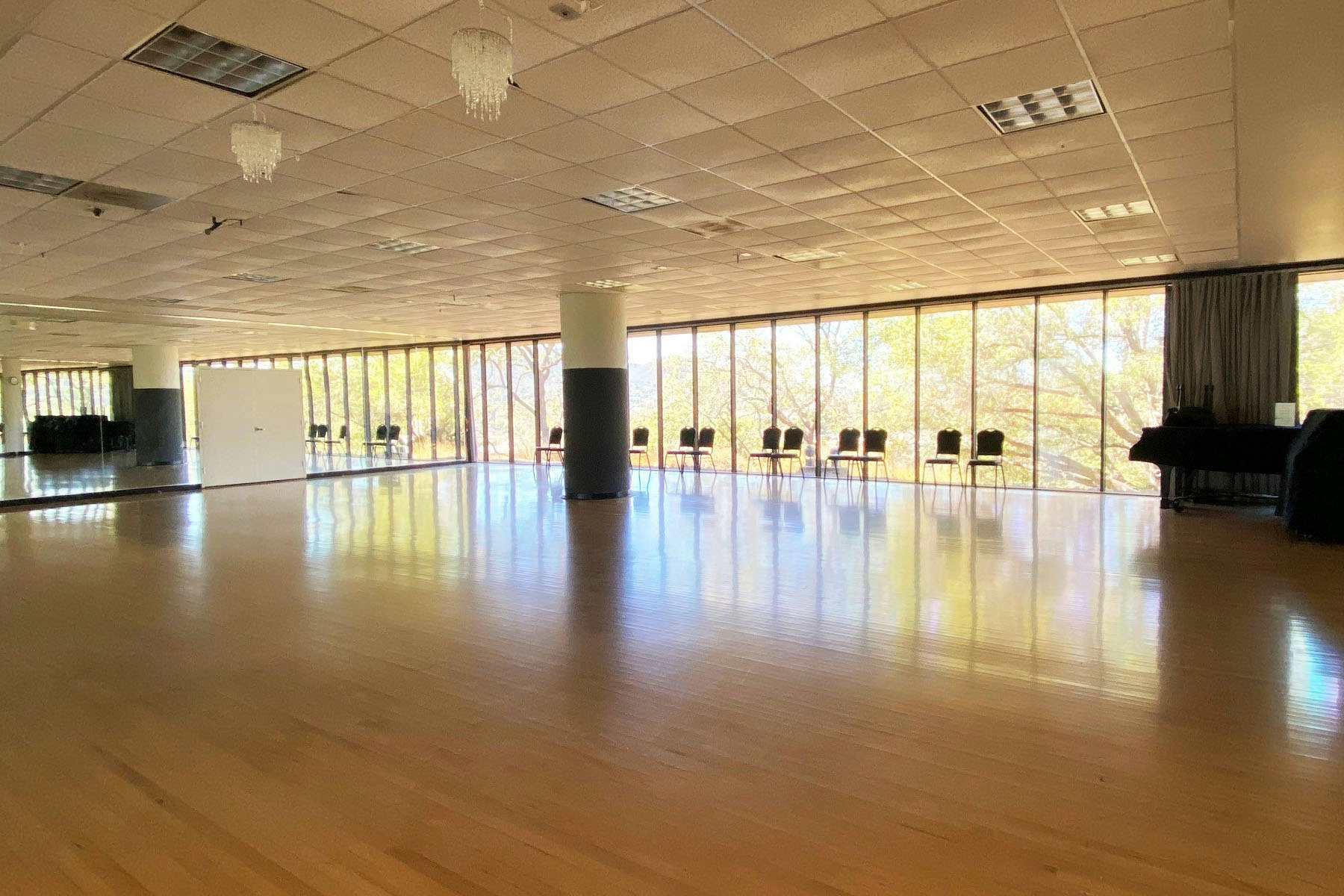 A spacious dance studio with wooden floors, large mirrors, chandeliers, and chairs lined up by the windows.
