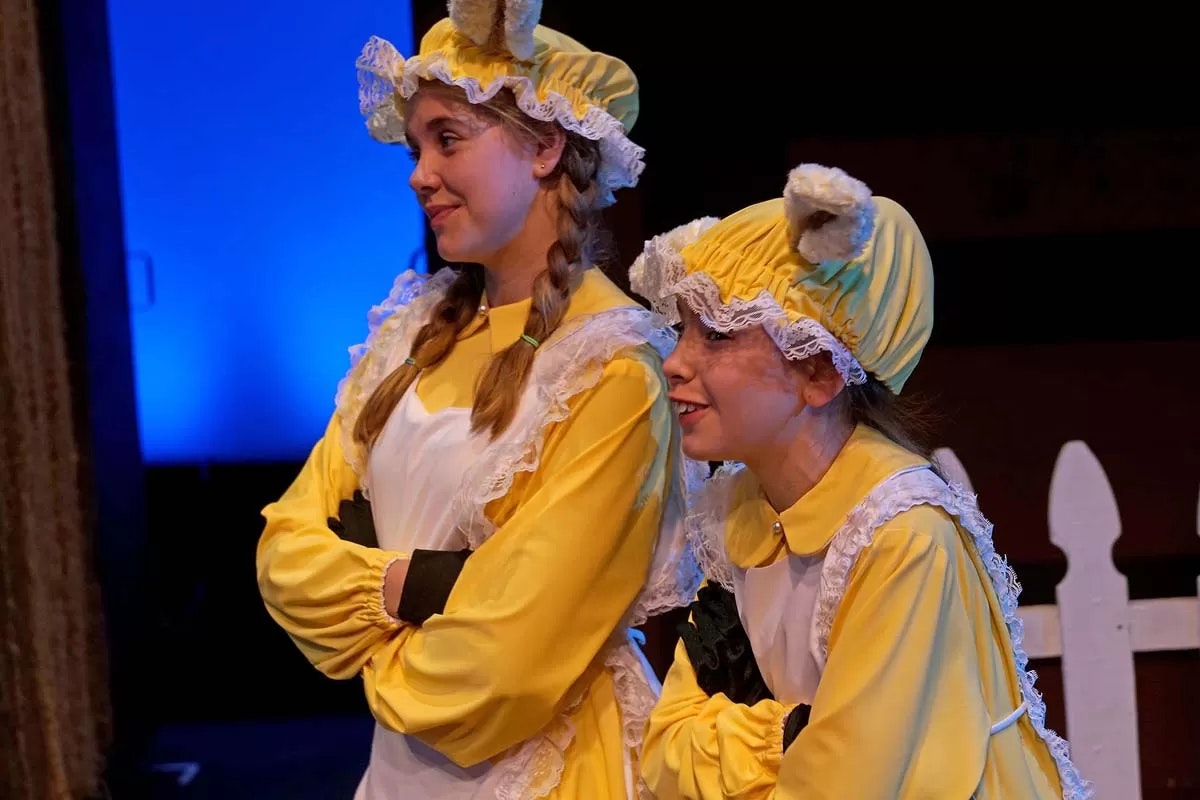 Two people in yellow and white costume dresses, with lace bonnets and gloves, on stage.