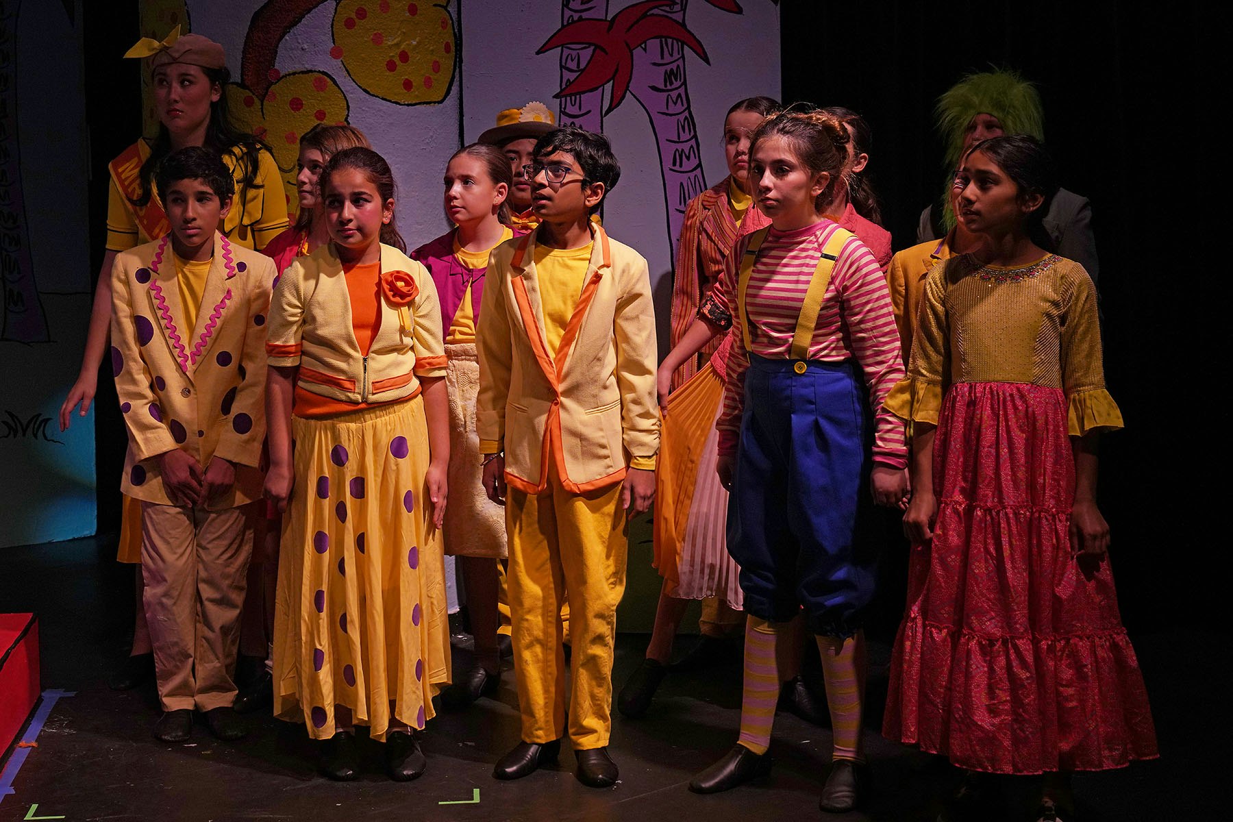A group of children in colorful costumes are performing on stage, featuring playful characters and a vibrant backdrop.
