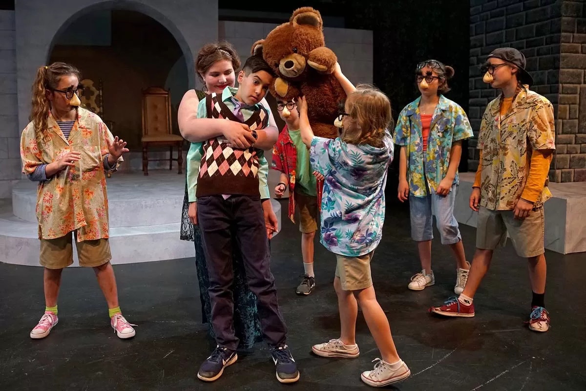 Group of kids on stage, wearing costumes, with funny glasses and noses; one holds a large teddy bear above their head.