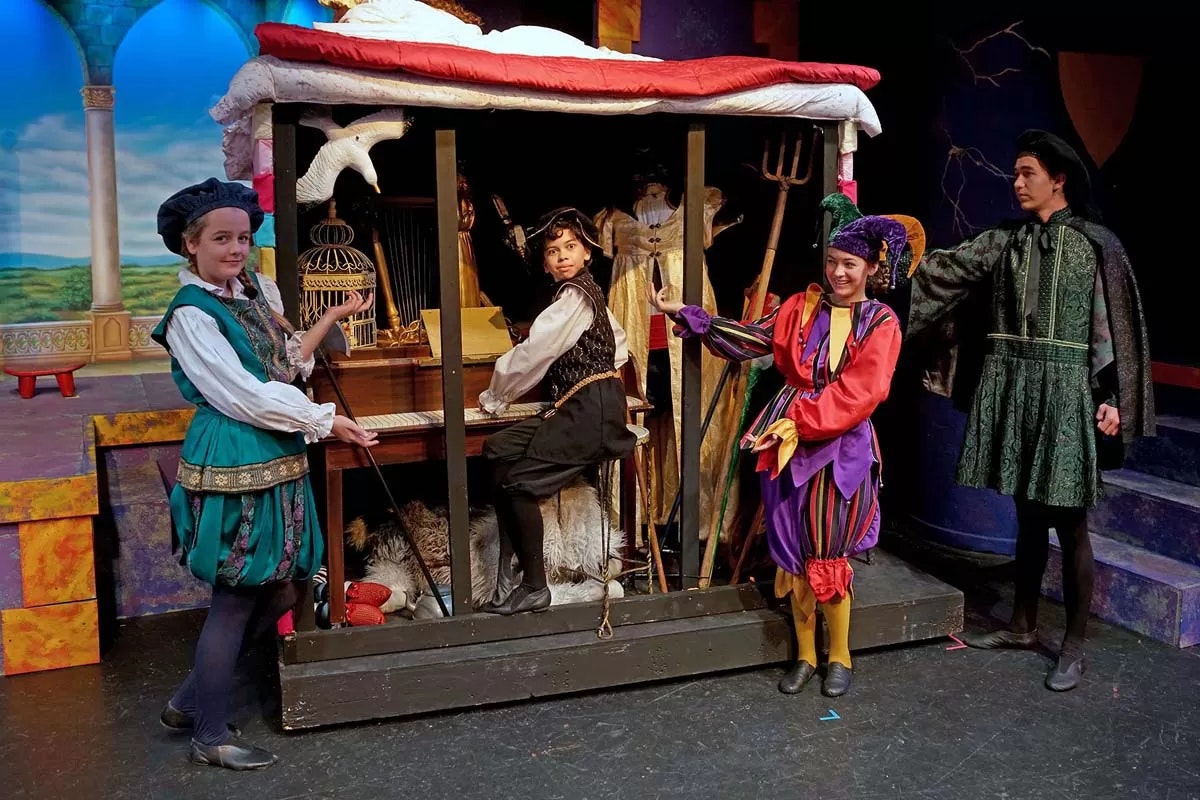 People in colorful costumes acting out a scene with props, including a birdcage and pitchfork, on a stage.