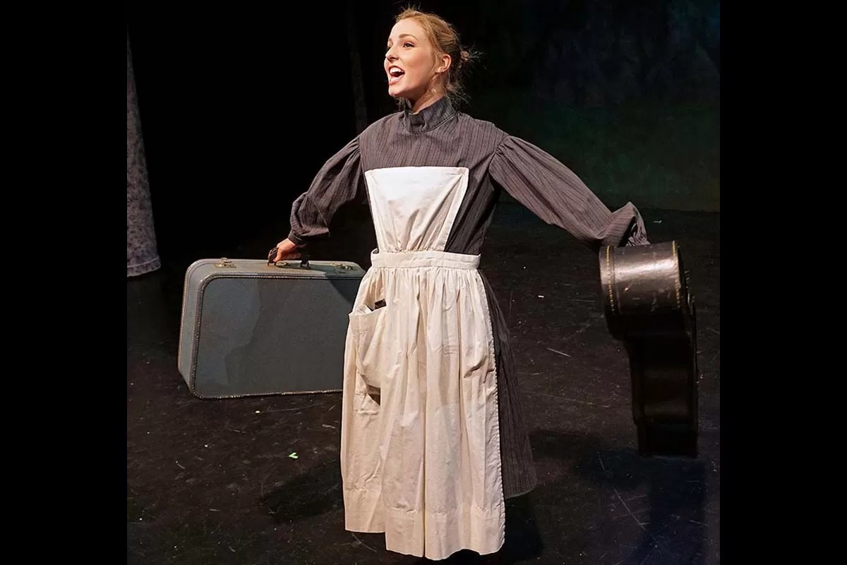 A person on stage in period costume holds a suitcase and a small case, with an expressive pose.