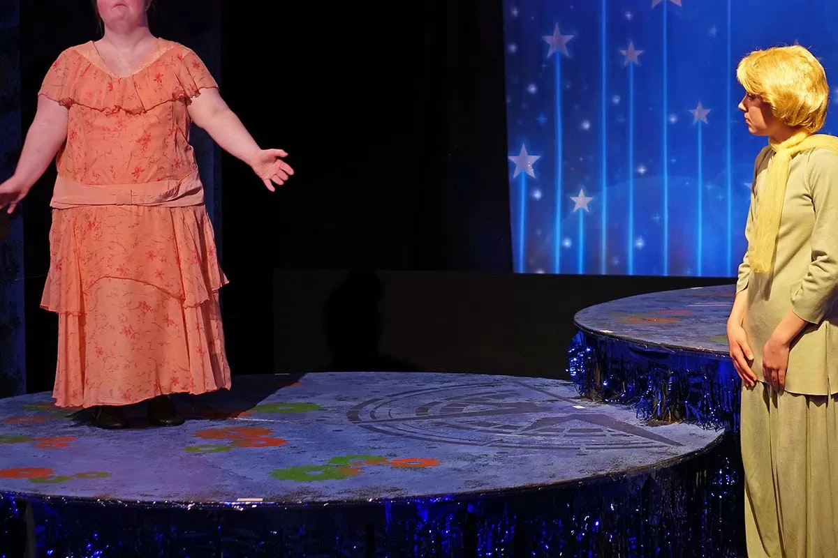 Two people on a stage, one in an orange dress with arms out, the other in costume looking on, starry backdrop.