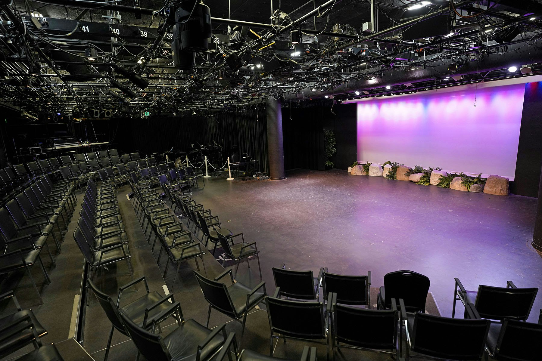Empty theater with black chairs, a large stage, overhead lighting equipment, and a backdrop with purple lighting.