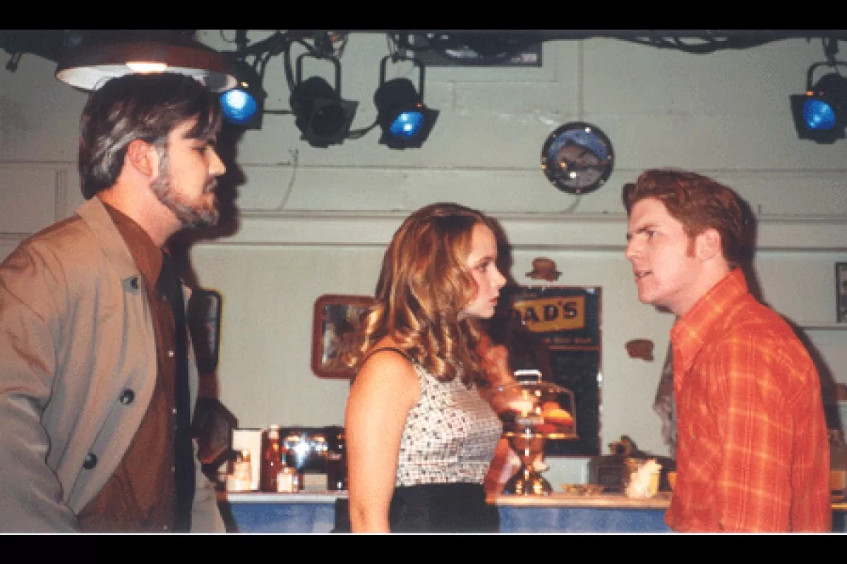 Three people are in a heated discussion at a bar with stage lights above them.