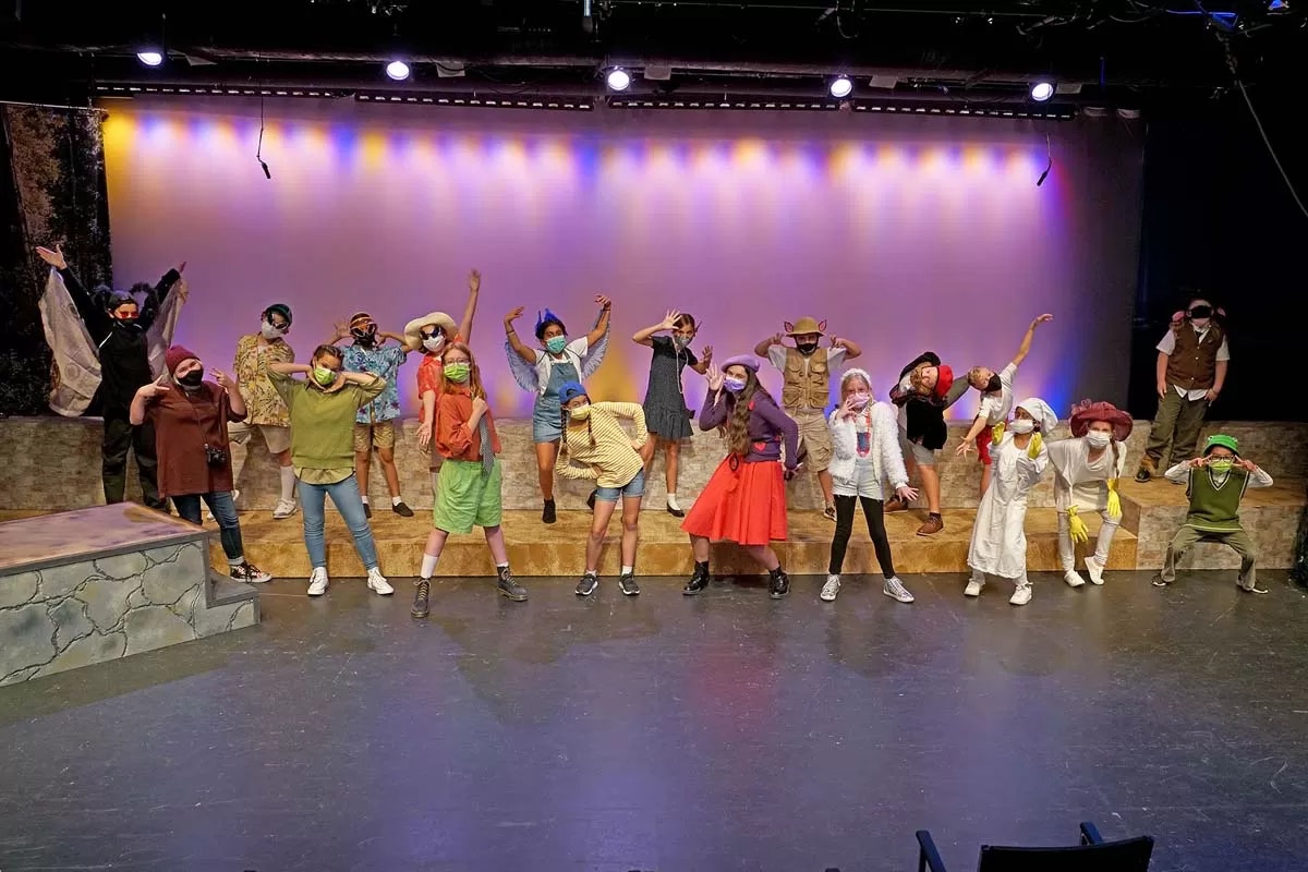 A group of people in colorful costumes performing on stage with a lit backdrop, posing energetically.