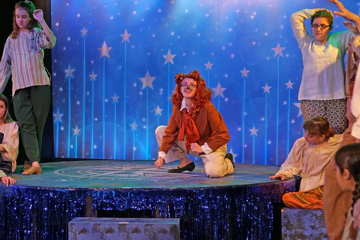 People in costume on stage, with a starry backdrop. A character in center crouches, surrounded by others.