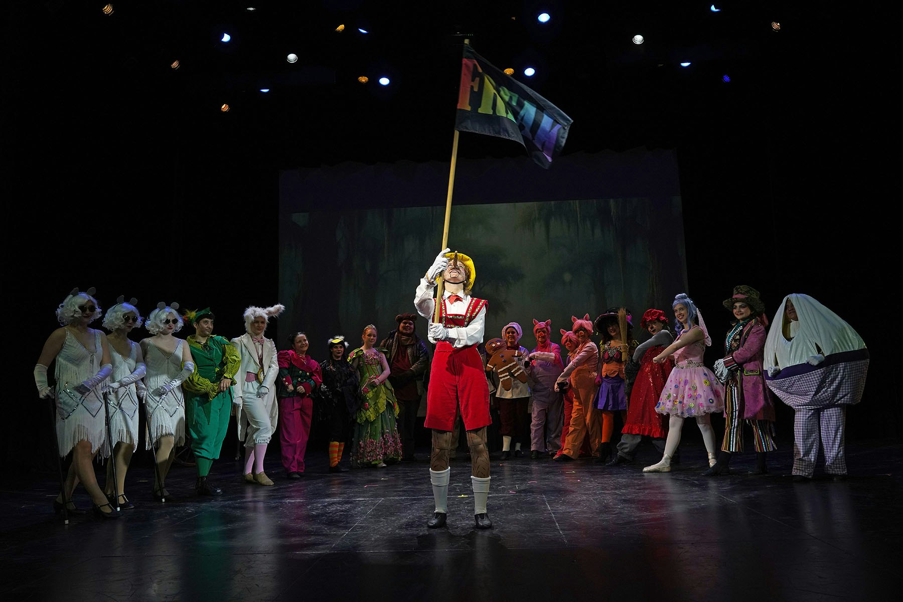 A theatrical performance with characters in colorful costumes on stage, one holding a “FREAK” flag.