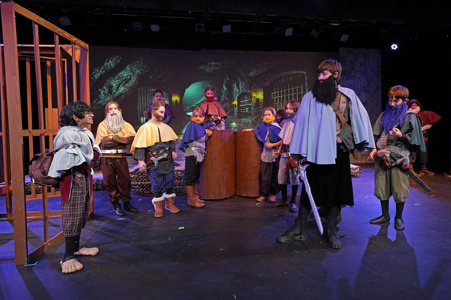 Children in medieval costumes with beards, swords, and props, on a stage with a fantasy cave backdrop.