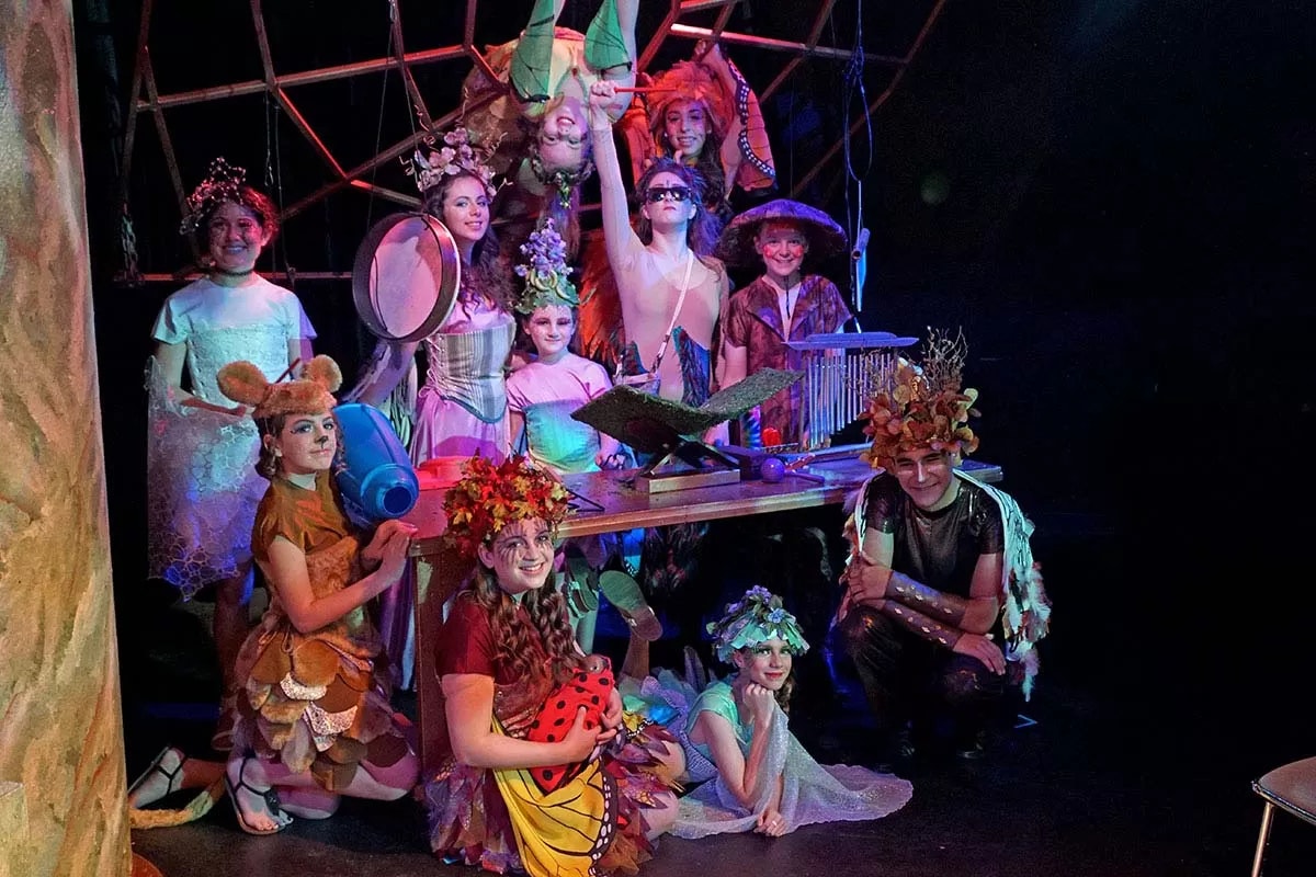 A group of people in colorful, nature-themed costumes posing on stage, some with floral and leaf designs, one upside down.