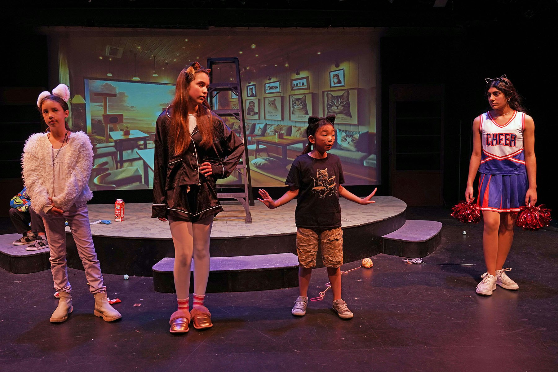 Four kids in costume on stage: one in a furry outfit, one in pajamas, one in a cat shirt, and one as a cheerleader.
