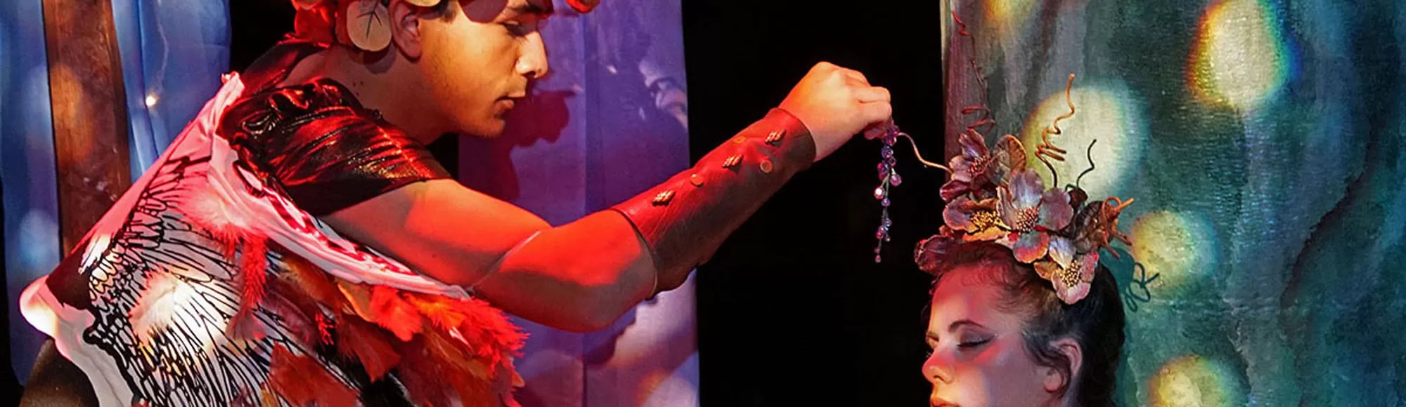 Two people in elaborate costumes; one holds a beaded object, the other with a floral headpiece. The scene is theatrical and colorful.