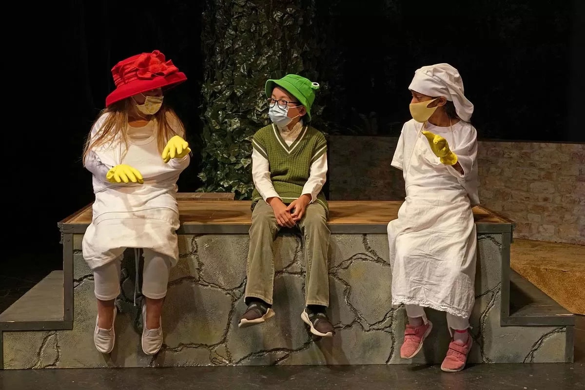 Three children in costume are sitting on stage; two wear white outfits with hats, and one wears green with glasses.