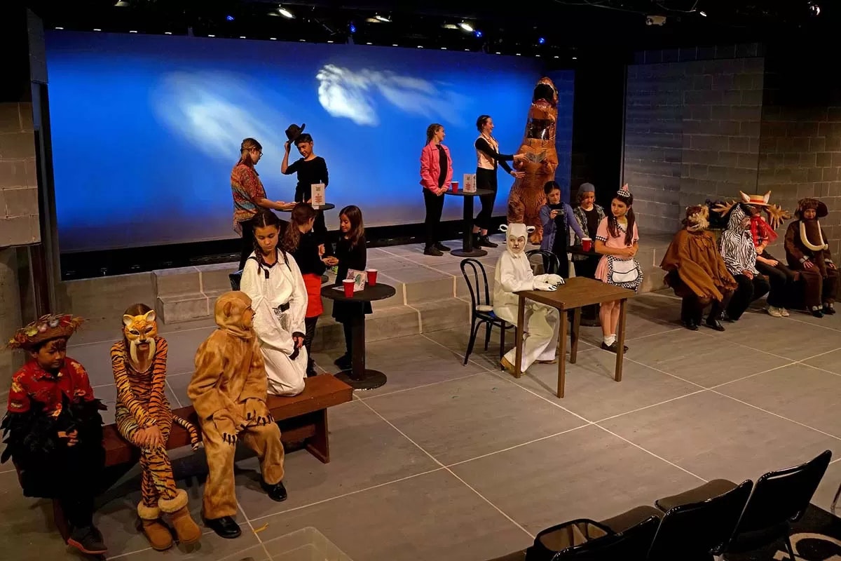 Kids on stage in animal costumes, a waitstaff and others interacting, set against a bright backdrop in a theater setting.