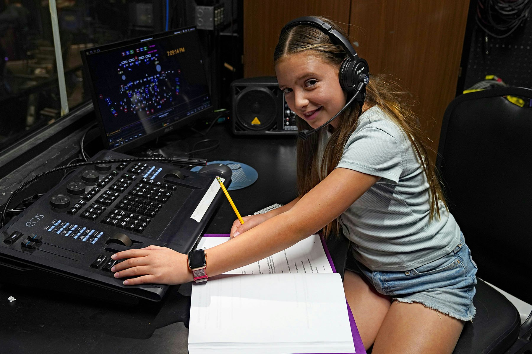 A young girl with headphones operates a control panel while writing notes in a studio setting, showcasing her involvement in production.