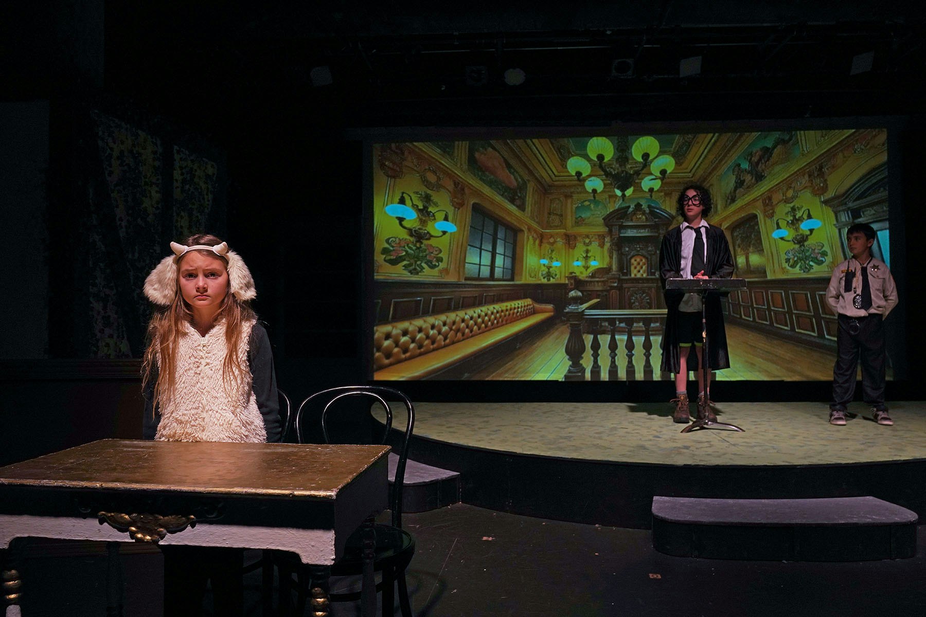 A stage scene with a child in a sheep costume, two others dressed as a judge and police officer, with a courtroom backdrop.