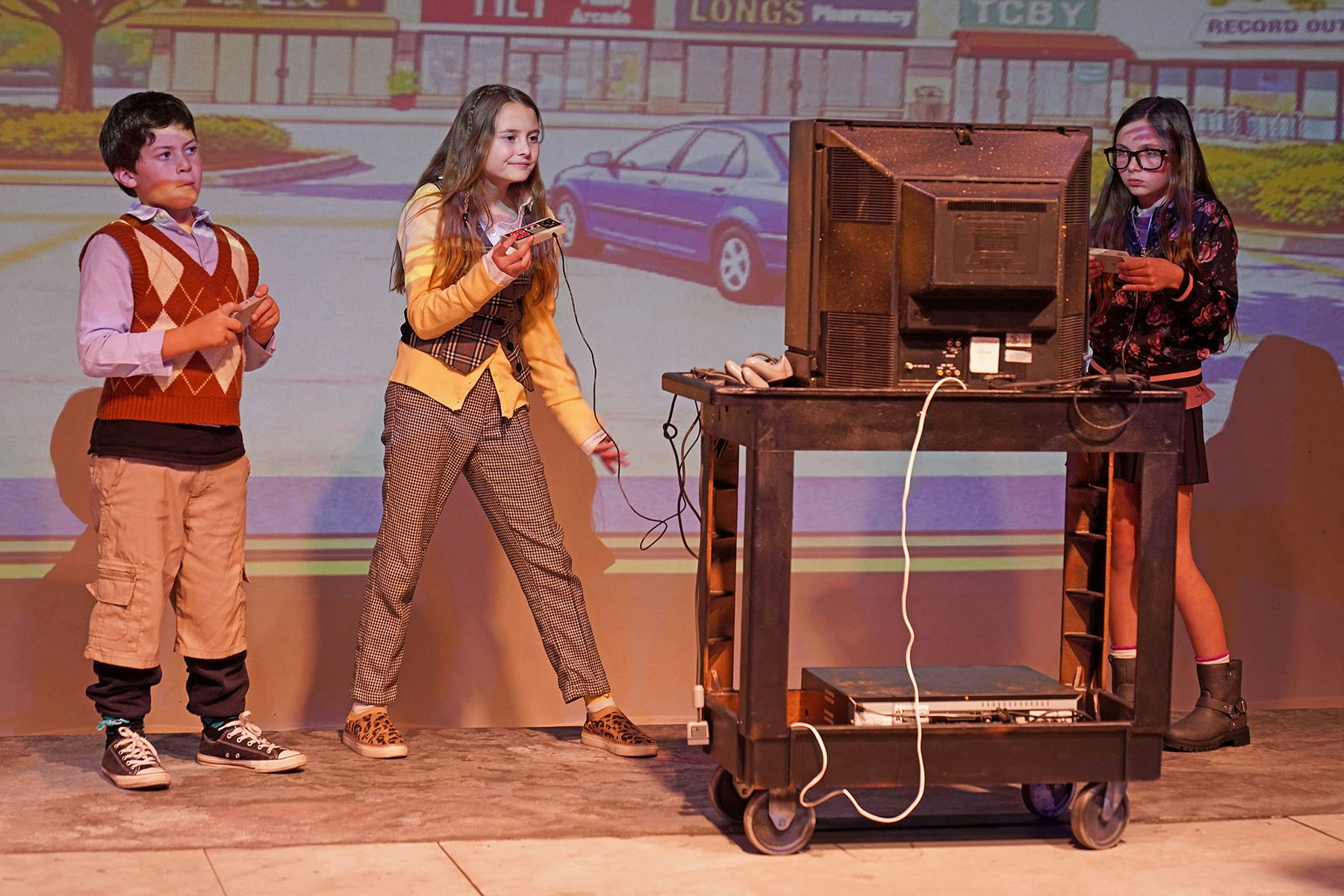 Three kids are interacting with a retro video game setup on stage, with a projection of a colorful arcade scene in the background.