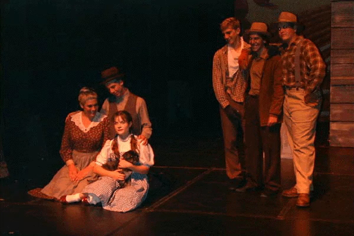 A theatrical scene with people in vintage clothing. A woman sits holding a small dog, surrounded by others standing and kneeling.