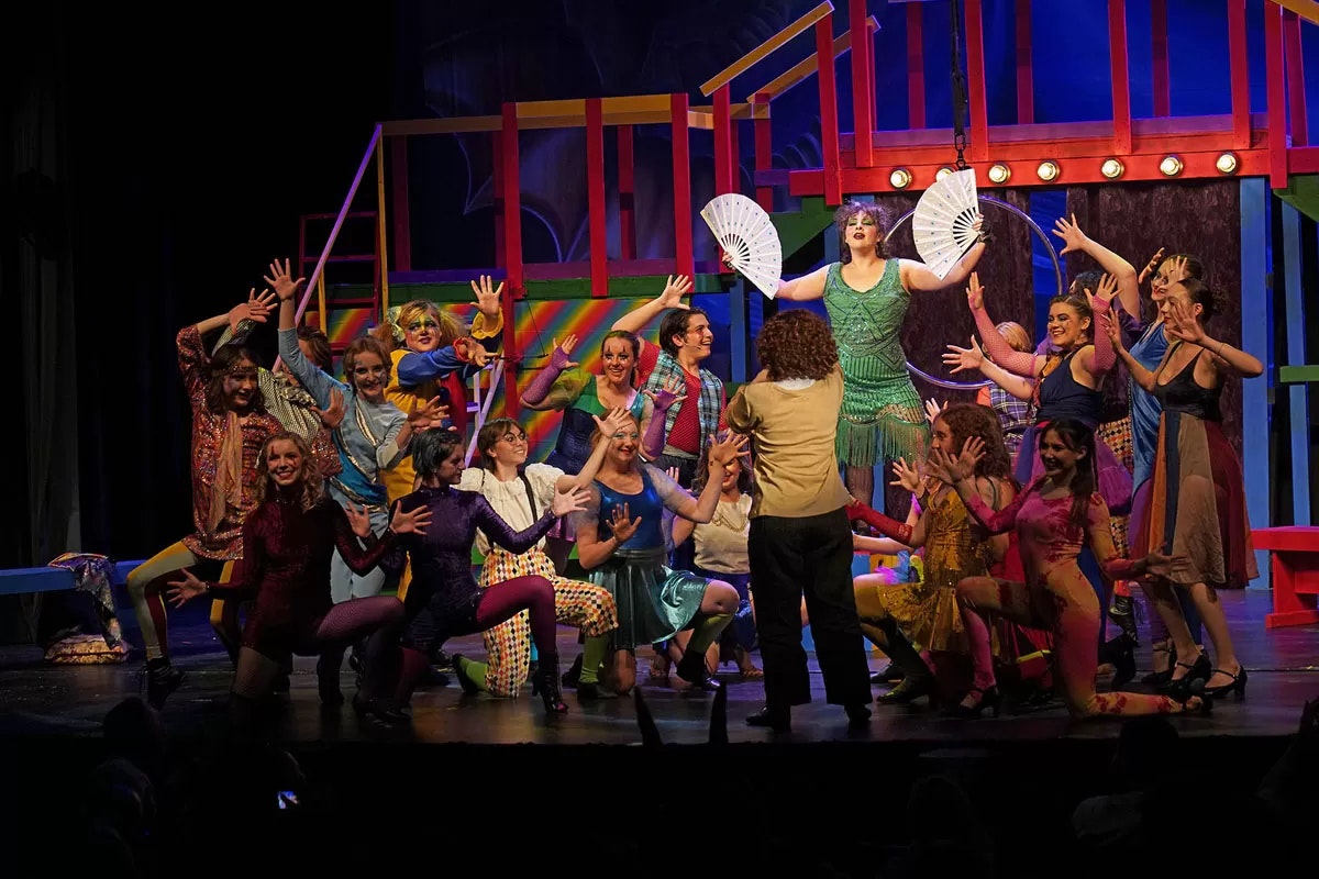 A large group of performers in colorful costumes pose energetically on stage with bright, theatrical lighting.