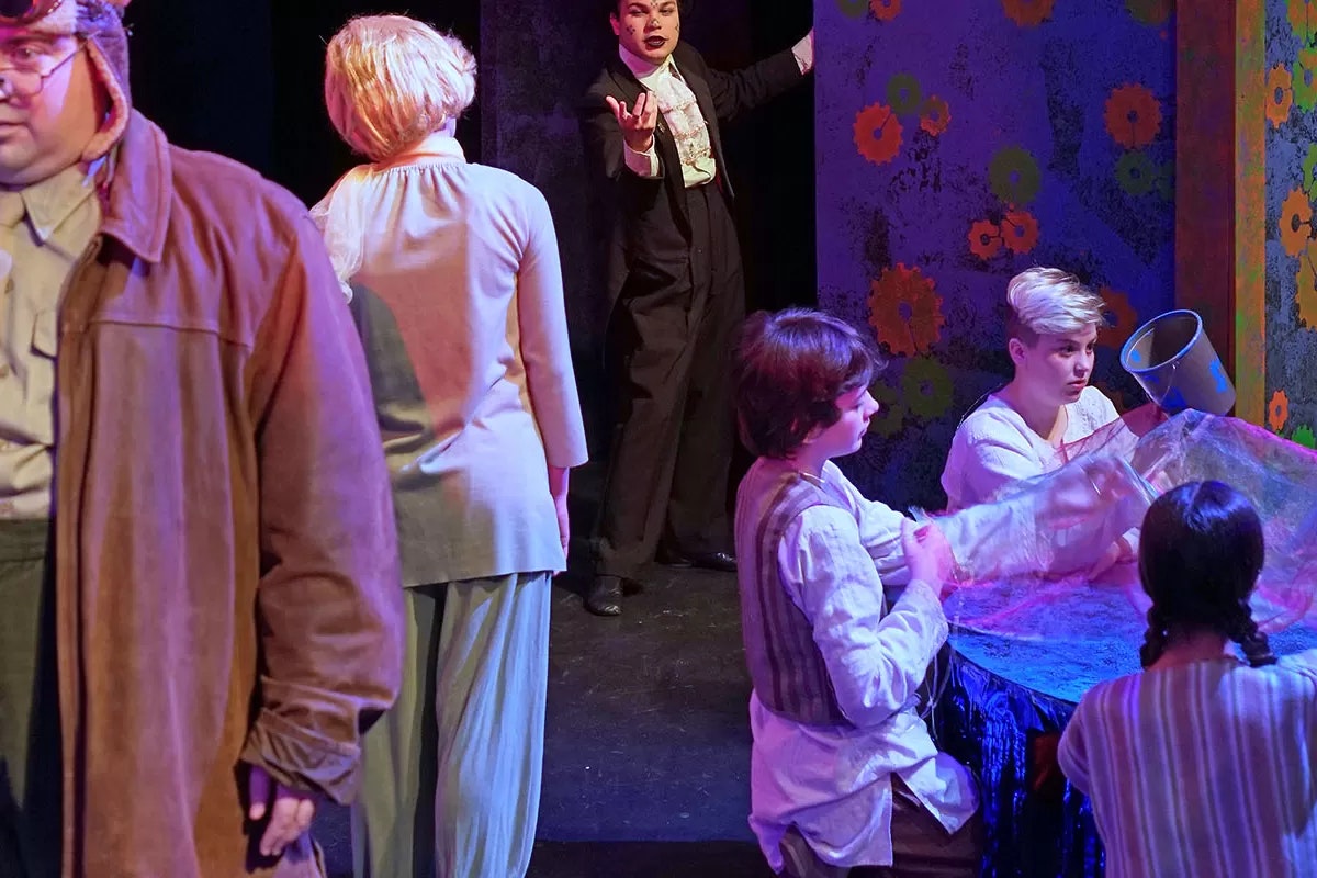 A theatrical scene with actors in vintage costumes, one gesturing dramatically, others gathered around a table with props.