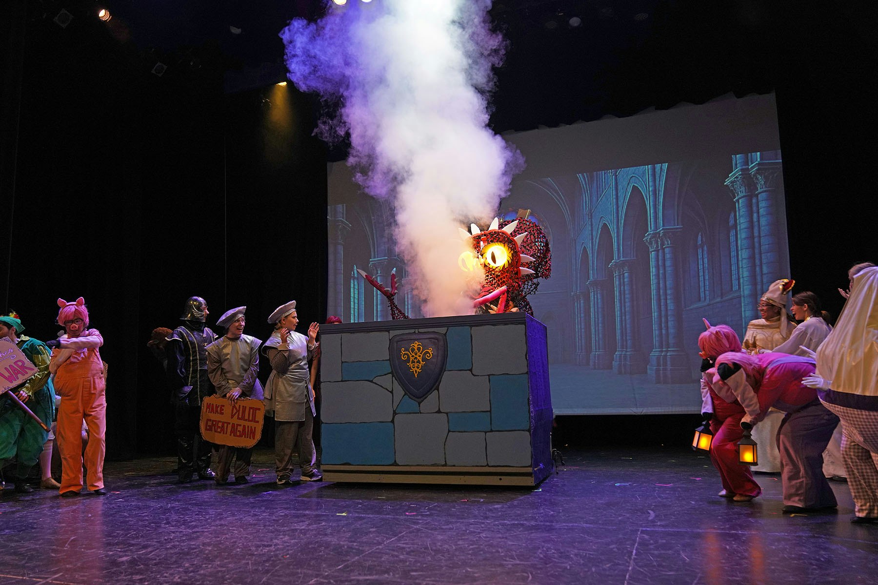 A theatrical scene with costumed characters, a dragon-like creature spewing smoke from a box, and a castle backdrop.