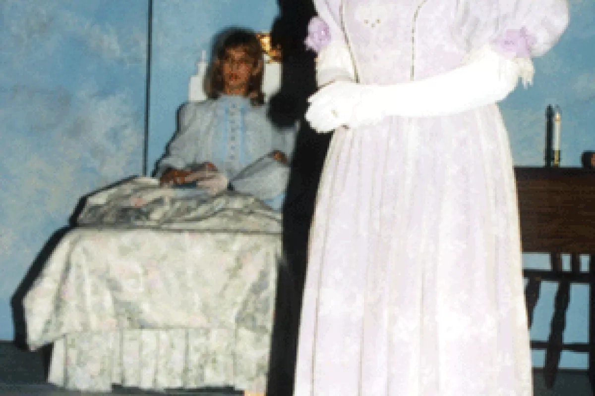 A person in historical attire stands near someone sitting in bed, set against a theatrical backdrop.