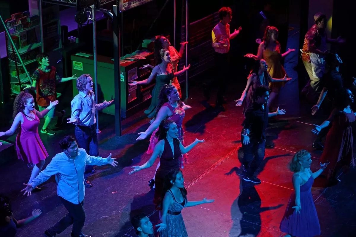 A group of people on stage performing a dance in colorful lighting, all with arms extended.