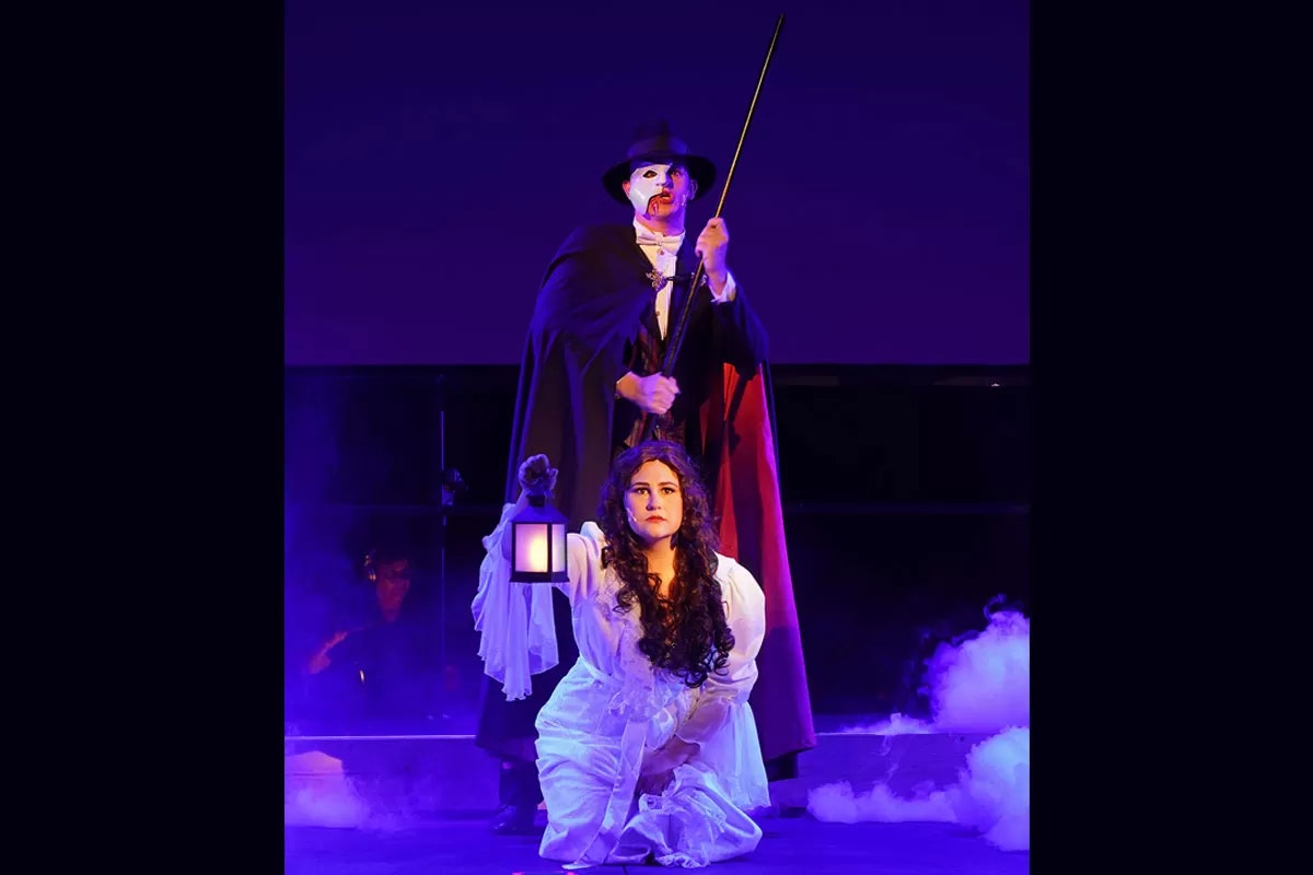 A masked figure in a cape stands with a sword; a kneeling woman holds a lantern amidst theatrical smoke.