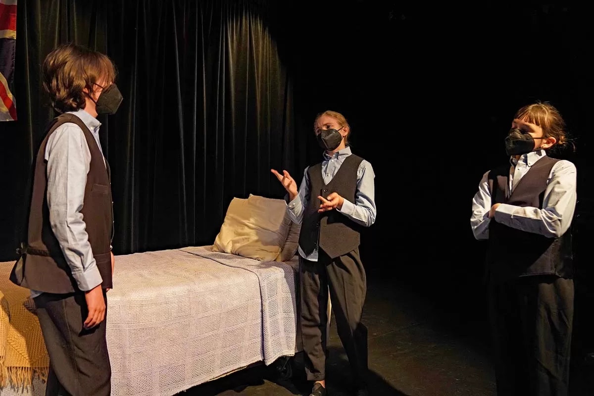 Three masked people in matching outfits are having a discussion on stage next to a bed.