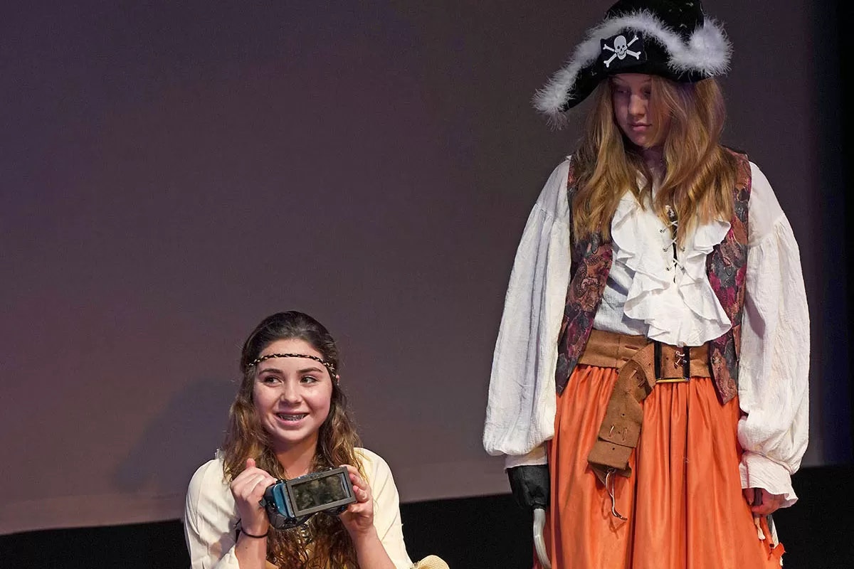 Two people in costumes; one holds a device and smiles, the other dressed as a pirate with a hat.
