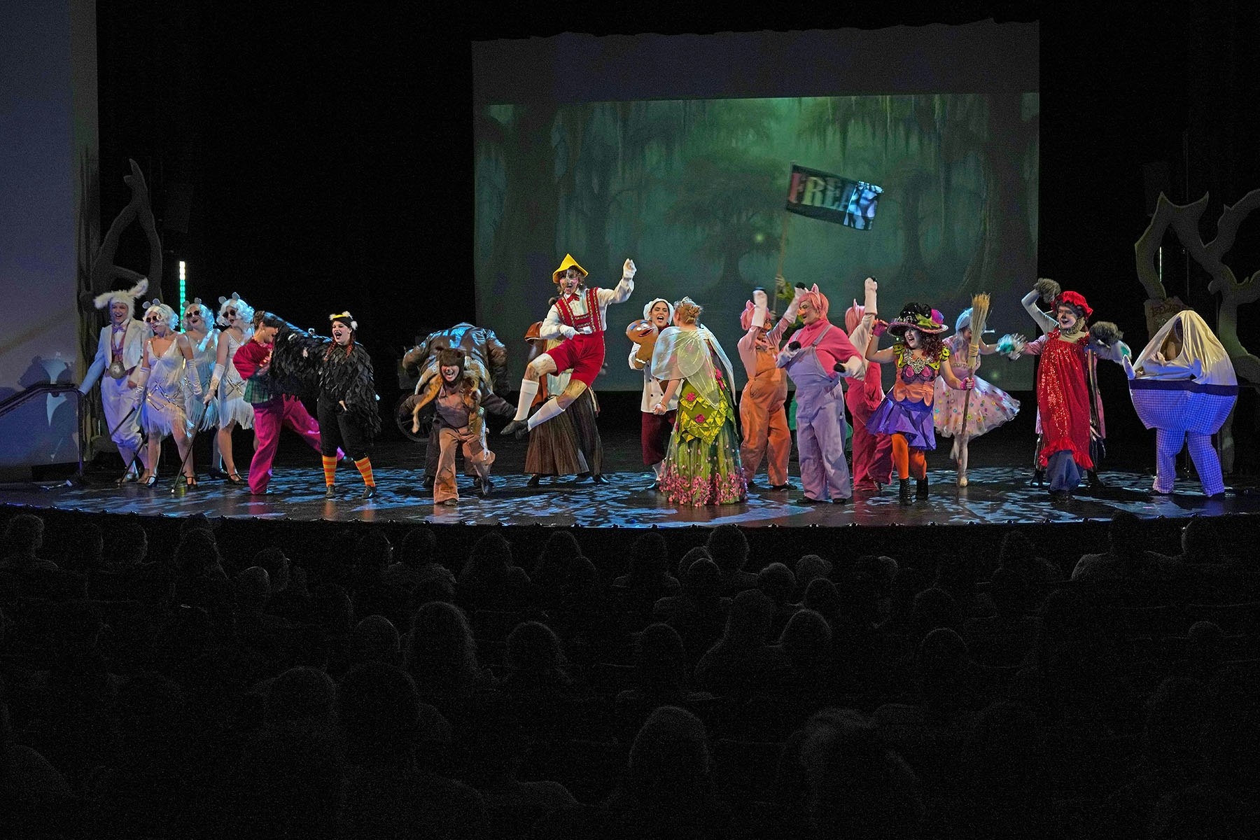 A group of performers in colorful, whimsical costumes on stage with a forest backdrop, entertaining an audience.