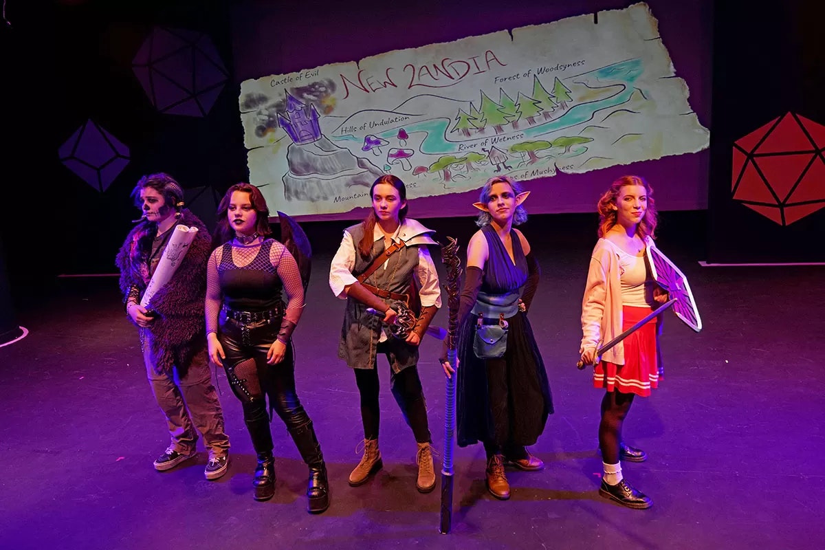 Five people in fantasy costumes on stage, with a map labeled "New Landia" in the background.