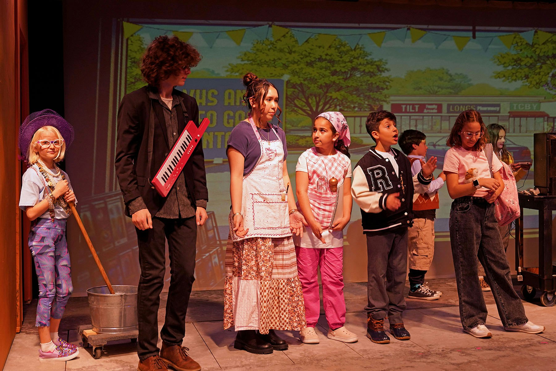 A group of kids in costumes performing on stage, showcasing various characters and props, with a colorful backdrop.