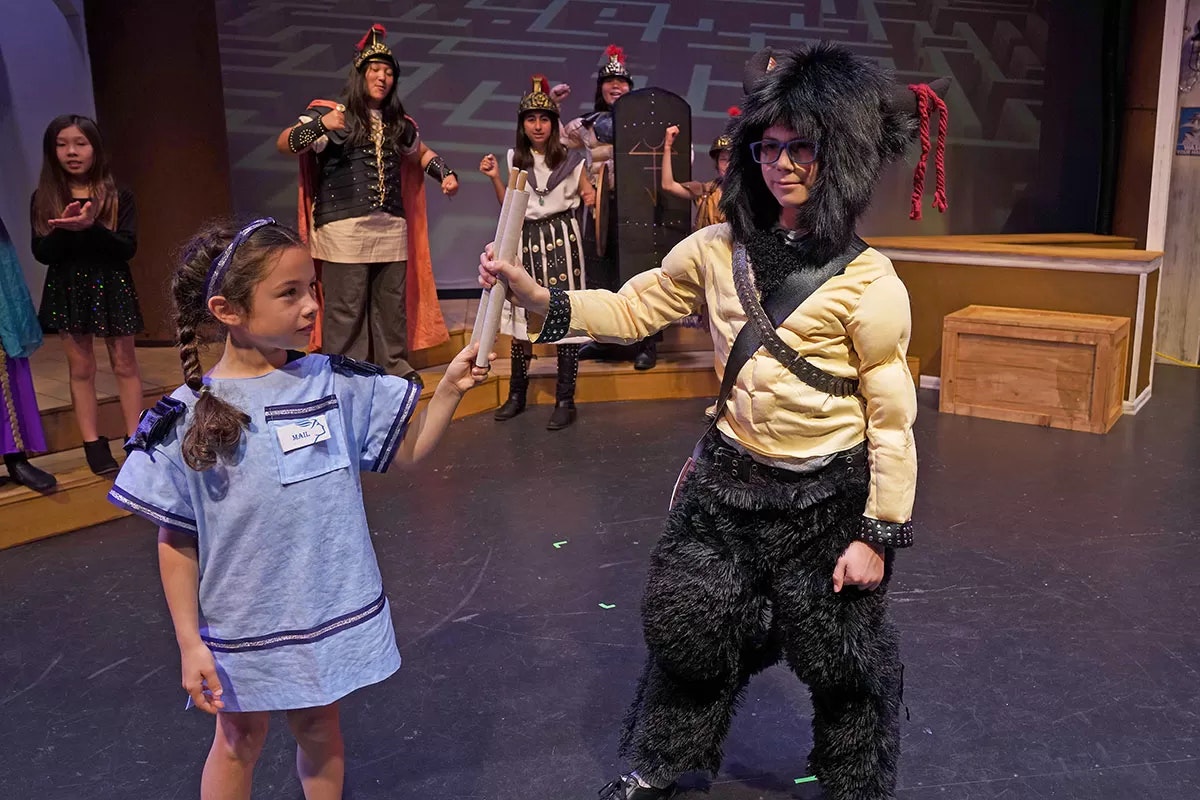 A child in a costume hands a scroll to another in a furry costume; others dressed as warriors stand in the background on stage.