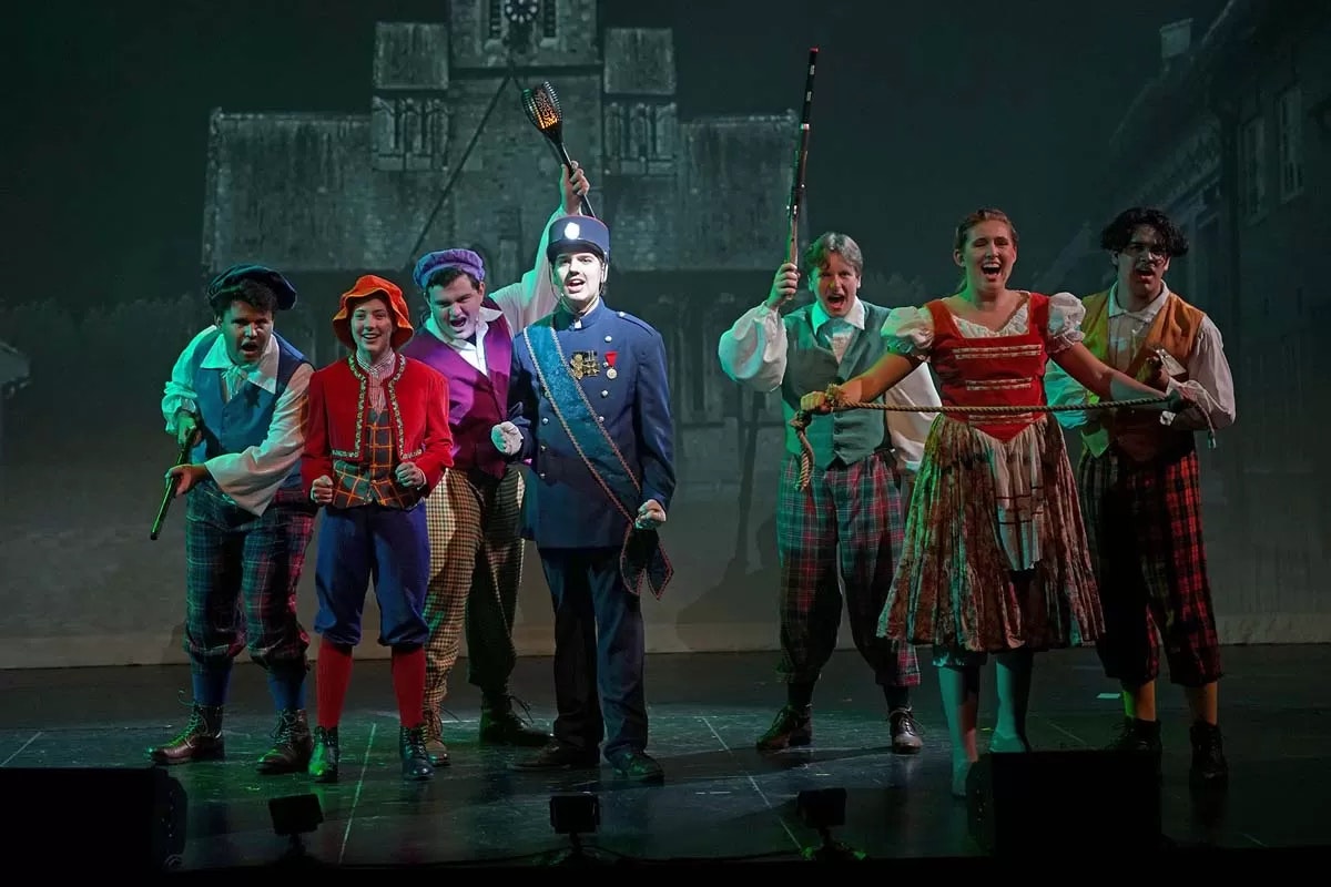 A group of performers in colorful costumes on stage, posing energetically with props against a backdrop.