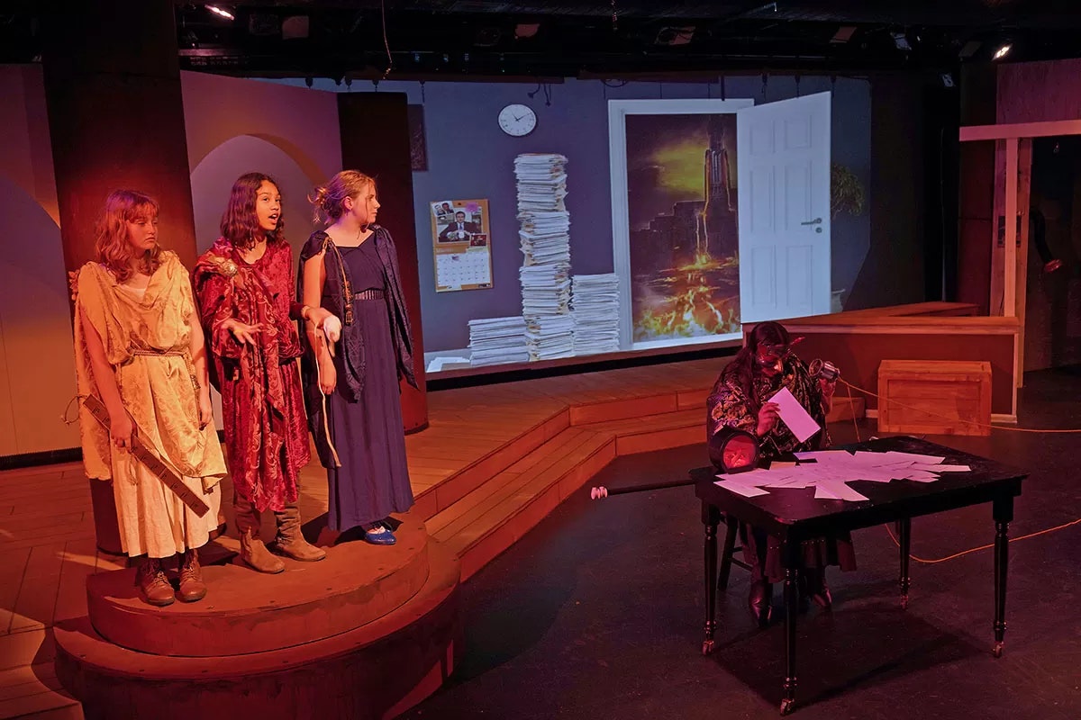 A theatrical scene with three people standing and one sitting at a table with papers, in front of a projected background.