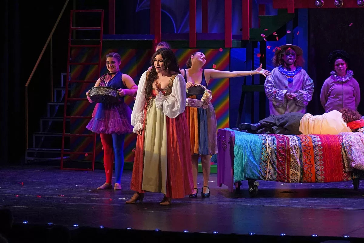 A colorful stage performance with actors in vibrant costumes, a rainbow backdrop, and a person lying on a decorated bed.