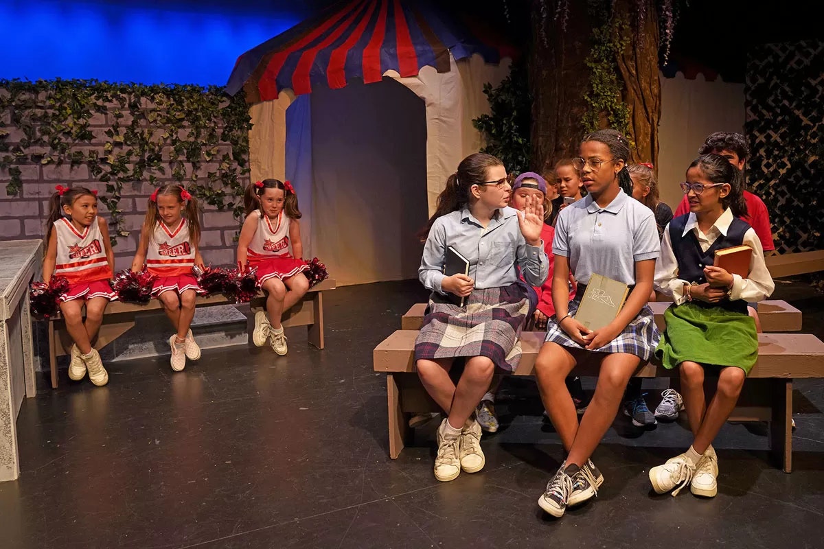 A group of children in a stage play; cheerleaders on the left, other children seated with books on the right, under a tent backdrop.